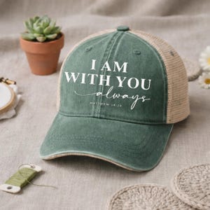 May include: A green baseball cap with a mesh back and the text "I AM WITH YOU always Matthew 28:20" in white. The cap has a distressed look and a curved brim. The background includes a small potted plant and crochet coasters.