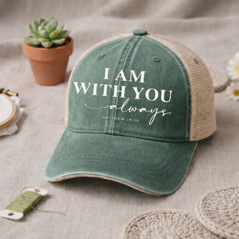 May include: A green baseball cap with a mesh back and the text "I AM WITH YOU always Matthew 28:20" in white. The cap has a distressed look and a curved brim. The background includes a small potted plant and crochet coasters.
