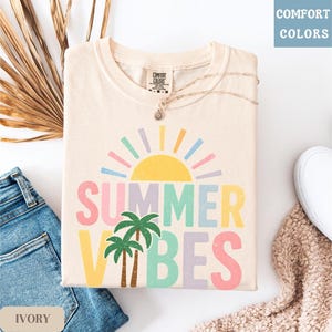 K&ouml;nnte beinhalten: Elfenbeinfarbenes Comfort Colors T-Shirt mit einem farbenfrohen "SUMMER VIBES"-Aufdruck. Das Design zeigt eine gelbe Sonne, Palmen und pastellfarbenen Text. Eine goldene Halskette liegt auf dem Shirt. Das Shirt liegt auf einer wei&szlig;en Oberfl&auml;che.