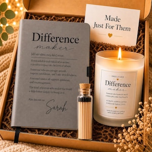 May include: A gift box containing a gray notebook with the text Difference maker and the name Sarah, a lit candle in a frosted glass jar with a label reading Smells Like Difference maker, and a small glass bottle filled with wooden matches.