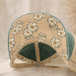 May include: A baseball cap with a floral daisy pattern on the underside of the brim. The cap features a green bill and a tan mesh back. The daisy pattern is white and yellow on a tan background.