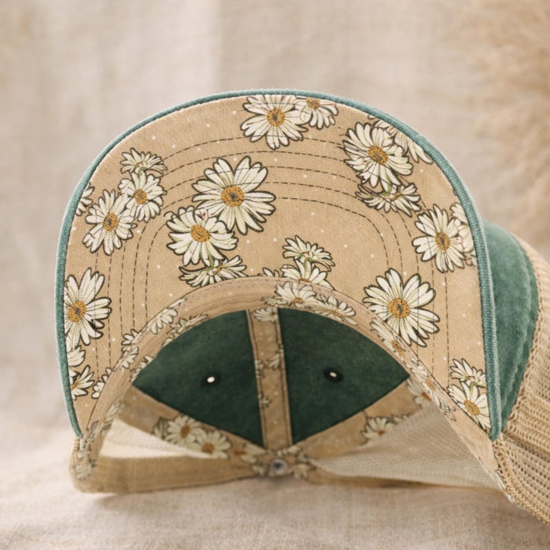 May include: A baseball cap with a floral daisy pattern on the underside of the brim. The cap features a green bill and a tan mesh back. The daisy pattern is white and yellow on a tan background.