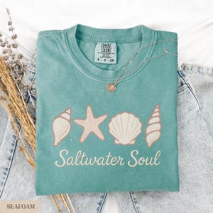 May include: Seafoam green t-shirt with a seashell, starfish, and shell graphic, and the words "Saltwater Soul". A silver necklace with a pendant rests on the shirt. The shirt is folded on top of denim jeans.