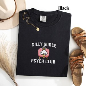 May include: Black t-shirt with a white embroidered design of a goose wearing glasses and sitting in a pink armchair. The text "Silly Goose Psych Club" is embroidered below the goose.