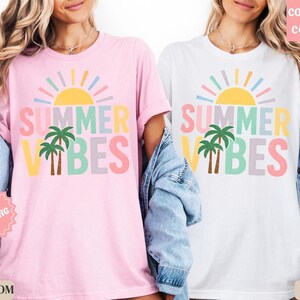 K&ouml;nnte beinhalten: Rosa und wei&szlig;e T-Shirts mit dem Text "SUMMER VIBES" in Pastellfarben, mit einer Sonne und Palmen-Grafik. Die Shirts sind Teil einer sommerlichen Bekleidungskollektion.