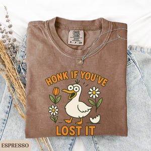 May include: Espresso-colored t-shirt with a cartoon duck graphic. The duck has wide eyes and is surrounded by flowers and the text "HONK IF YOU'VE LOST IT". A silver necklace rests on the shirt, which is placed on a pair of blue jeans.