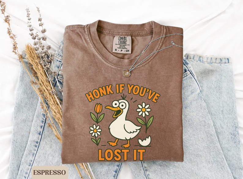 May include: Espresso-colored t-shirt with a cartoon duck graphic. The duck has wide eyes and is surrounded by flowers and the text "HONK IF YOU'VE LOST IT". A silver necklace rests on the shirt, which is placed on a pair of blue jeans.