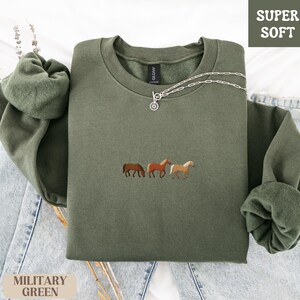 K&ouml;nnte beinhalten: Milit&auml;rgr&uuml;nes Sweatshirt mit Pferde-Stickerei. Der Rundhals-Pullover tr&auml;gt die Aufschrift "SUPER SOFT" in einem wei&szlig;en Feld. Eine silberne Halskette mit Anh&auml;nger liegt auf dem Sweatshirt. Das Sweatshirt liegt auf einer blauen Jeans.