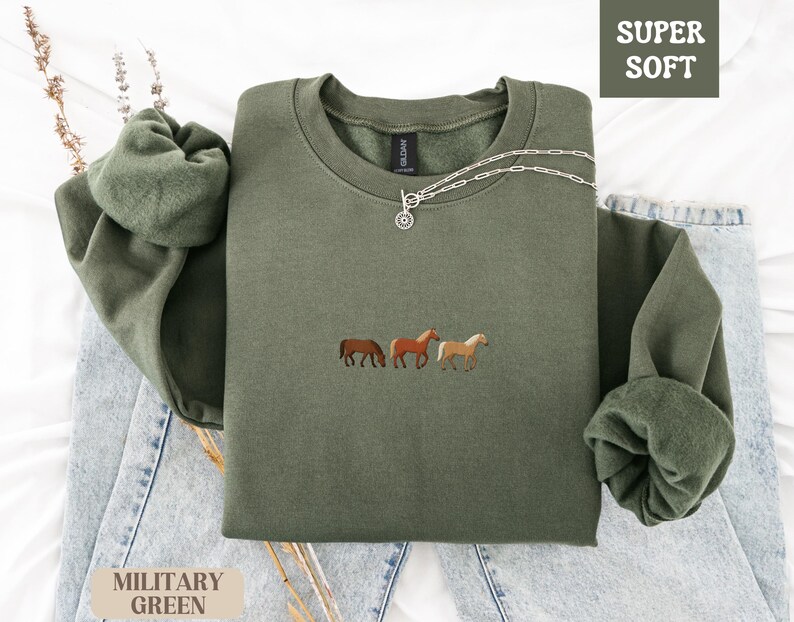 K&ouml;nnte beinhalten: Milit&auml;rgr&uuml;nes Sweatshirt mit Pferde-Stickerei. Der Rundhals-Pullover tr&auml;gt die Aufschrift "SUPER SOFT" in einem wei&szlig;en Feld. Eine silberne Halskette mit Anh&auml;nger liegt auf dem Sweatshirt. Das Sweatshirt liegt auf einer blauen Jeans.