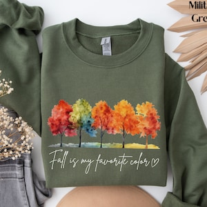 May include: A military green sweatshirt with a watercolor design of five trees with colorful leaves. The text "Fall is my favorite color" is printed below the trees with a heart.