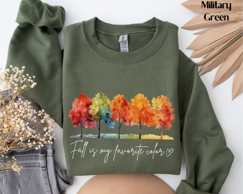 May include: A military green sweatshirt with a watercolor design of five trees with colorful leaves. The text "Fall is my favorite color" is printed below the trees with a heart.
