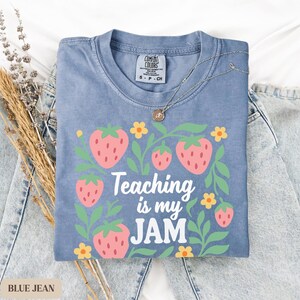 May include: A blue jean colored t-shirt with a strawberry graphic and the text "Teaching is my JAM". The shirt is paired with light blue jeans and a silver necklace. The shirt is a Comfort Colors brand.