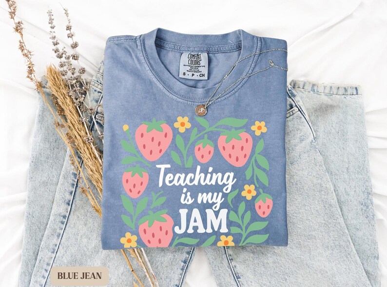May include: A blue jean colored t-shirt with a strawberry graphic and the text "Teaching is my JAM". The shirt is paired with light blue jeans and a silver necklace. The shirt is a Comfort Colors brand.