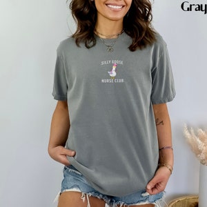 Puede incluir: Camiseta gris de manga corta con el texto "SILLY GOOSE NURSE CLUB" sobre un dibujo de un ganso con gorro de enfermera y un malet&iacute;n m&eacute;dico. La camiseta est&aacute; hecha de un material suave.