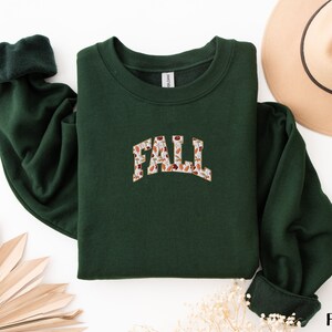 May include: A dark green sweatshirt with a white and brown "FALL" graphic featuring a floral pattern. The sweatshirt has a crew neck and long sleeves.