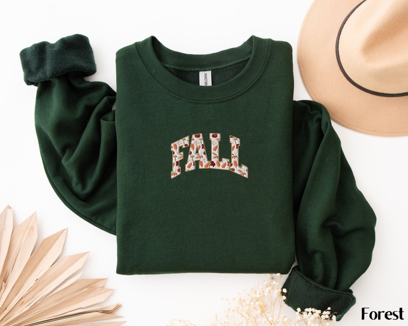 May include: A dark green sweatshirt with a white and brown "FALL" graphic featuring a floral pattern. The sweatshirt has a crew neck and long sleeves.