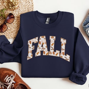 K&ouml;nnte beinhalten: Navyblauer Sweatshirt mit einem wei&szlig;en "FALL"-Motiv. Das Motiv besteht aus einem Muster aus Herbstbl&auml;ttern und Eicheln.
