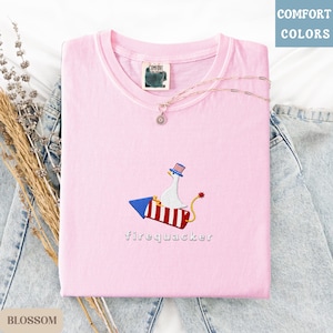 K&ouml;nnte beinhalten: Hellrosa T-Shirt mit gesticktem Design einer Gans auf einer Rakete, darunter das Wort "firequacker". Das T-Shirt hat einen Rundhalsausschnitt und ein "Comfort Colors"-Etikett. Eine zarte Halskette mit Anh&auml;nger liegt auf dem T-Shirt, das auf Denim gefaltet ist.