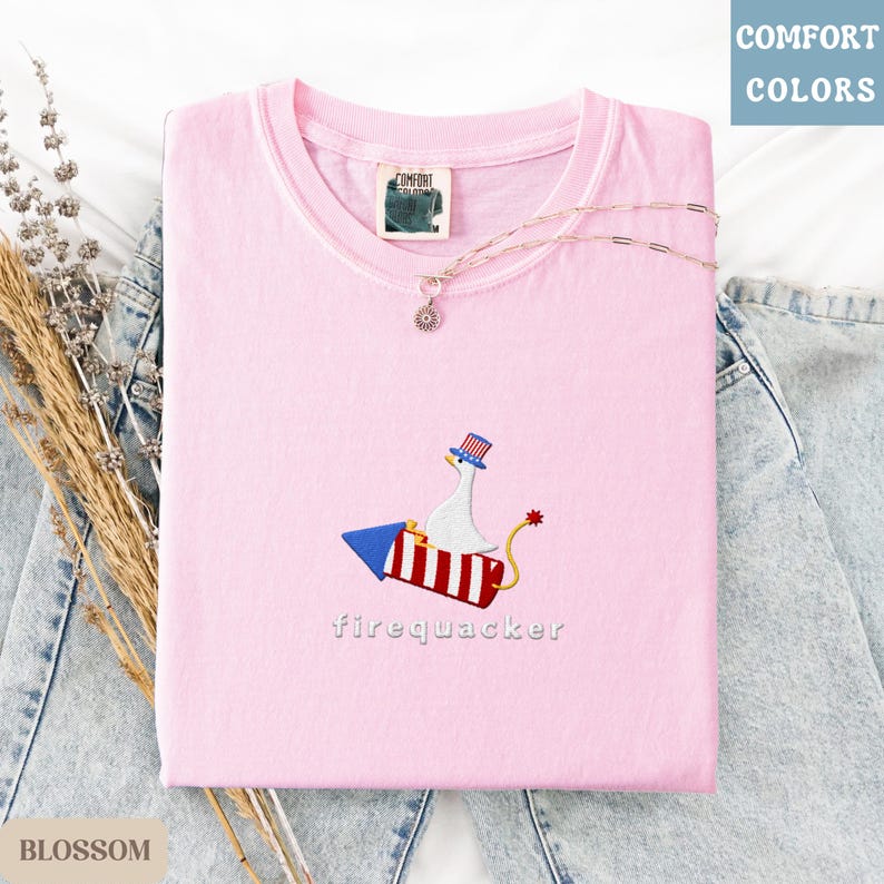 K&ouml;nnte beinhalten: Hellrosa T-Shirt mit gesticktem Design einer Gans auf einer Rakete, darunter das Wort "firequacker". Das T-Shirt hat einen Rundhalsausschnitt und ein "Comfort Colors"-Etikett. Eine zarte Halskette mit Anh&auml;nger liegt auf dem T-Shirt, das auf Denim gefaltet ist.