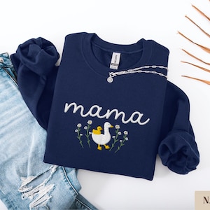 May include: Navy blue sweatshirt with the word "mama" embroidered in white with a white duck and a yellow duckling embroidered below. The duck and duckling are surrounded by small white flowers.