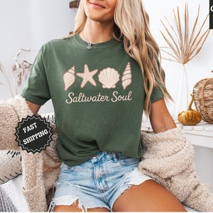May include: Moss green t-shirt with a seashell, starfish, and shell design, and the words "Saltwater Soul". The shirt is part of the Comfort Colors collection. The model is wearing denim shorts and a beige sweater.