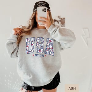 Peut inclure: Un sweat-shirt gris clair avec le mot "USA" en motif floral. Le sweat-shirt affiche &eacute;galement le texte "EST. 1776" et une &eacute;toile rouge. Le mod&egrave;le porte une casquette de baseball noire.
