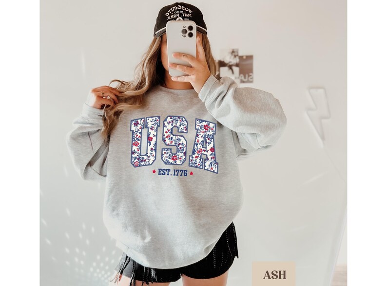 Peut inclure: Un sweat-shirt gris clair avec le mot "USA" en motif floral. Le sweat-shirt affiche &eacute;galement le texte "EST. 1776" et une &eacute;toile rouge. Le mod&egrave;le porte une casquette de baseball noire.