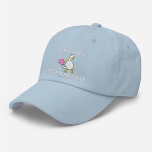 May include: A light blue baseball cap with the text "Silly Goose Pickleball Club" embroidered on the front. A cartoon goose holding a pink pickleball paddle is also embroidered on the cap.