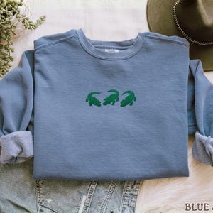 May include: A blue jean-colored sweatshirt with three green alligator designs embroidered across the chest. The sweatshirt has a crew neck and long sleeves with cuffed wrists. The text "BLUE JEAN" is printed at the bottom right.