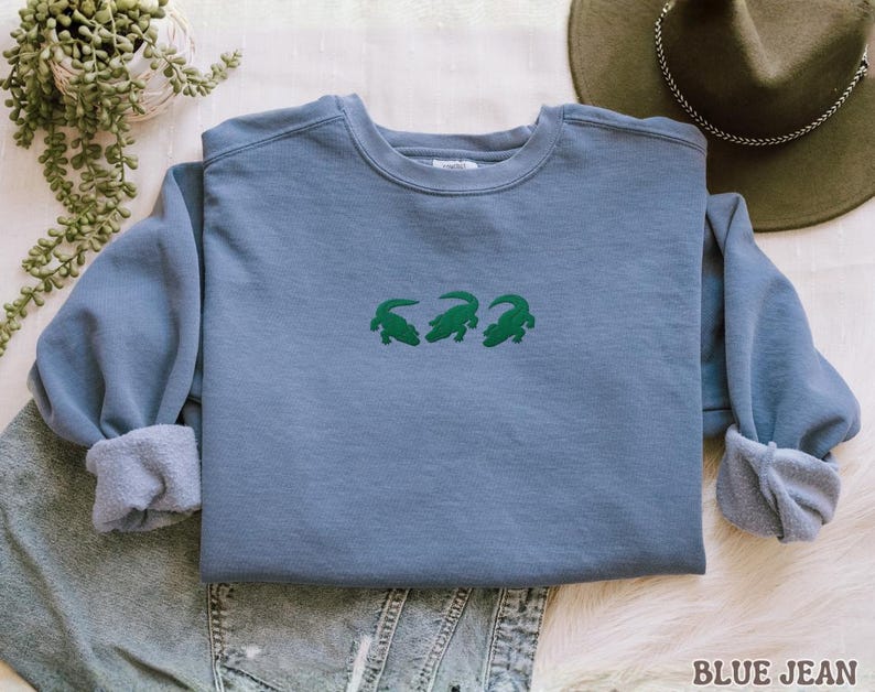 May include: A blue jean-colored sweatshirt with three green alligator designs embroidered across the chest. The sweatshirt has a crew neck and long sleeves with cuffed wrists. The text "BLUE JEAN" is printed at the bottom right.
