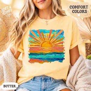 Puede incluir: Camiseta amarilla clara con un dise&ntilde;o de puesta de sol colorido. El gr&aacute;fico presenta un sol vibrante con rayos naranjas sobre un horizonte azul y naranja. La camiseta tiene las palabras "Comfort Colors" y "Soft & Comfy".
