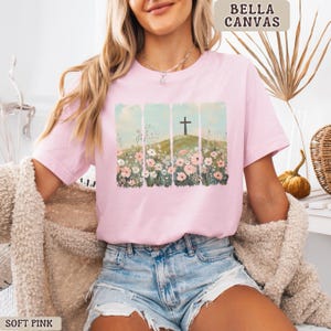 May include: Soft pink Bella Canvas t-shirt featuring a graphic of a cross on a hill surrounded by flowers. The design is set against a backdrop of vertical light blue panels. The shirt is a soft pink color.