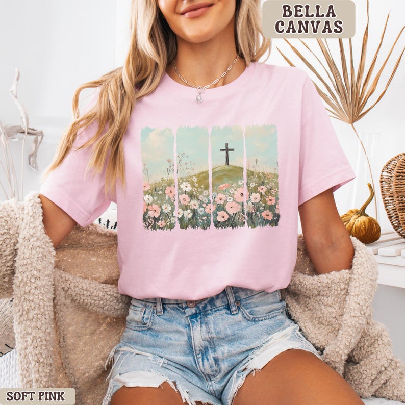 May include: Soft pink Bella Canvas t-shirt featuring a graphic of a cross on a hill surrounded by flowers. The design is set against a backdrop of vertical light blue panels. The shirt is a soft pink color.