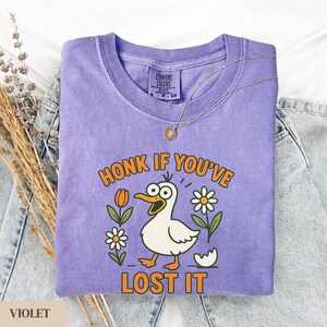 May include: Violet Comfort Colors t-shirt with a cartoon duck graphic. The duck has an open beak and is surrounded by flowers. The text "HONK IF YOU'VE LOST IT" is printed in orange. A gold necklace is draped over the shirt.