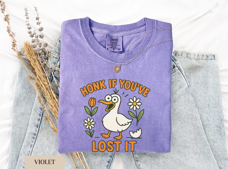May include: Violet Comfort Colors t-shirt with a cartoon duck graphic. The duck has an open beak and is surrounded by flowers. The text "HONK IF YOU'VE LOST IT" is printed in orange. A gold necklace is draped over the shirt.