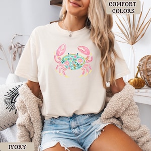K&ouml;nnte beinhalten: Elfenbeinfarbenes T-Shirt mit einem Aquarell-Krebs-Design. Der Krebs hat rosa Scheren, einen t&uuml;rkisfarbenen K&ouml;rper voller Blumen und gelbe Akzente. Die Worte "Comfort Colors" und "Ivory" sind auf dem Bild zu sehen.
