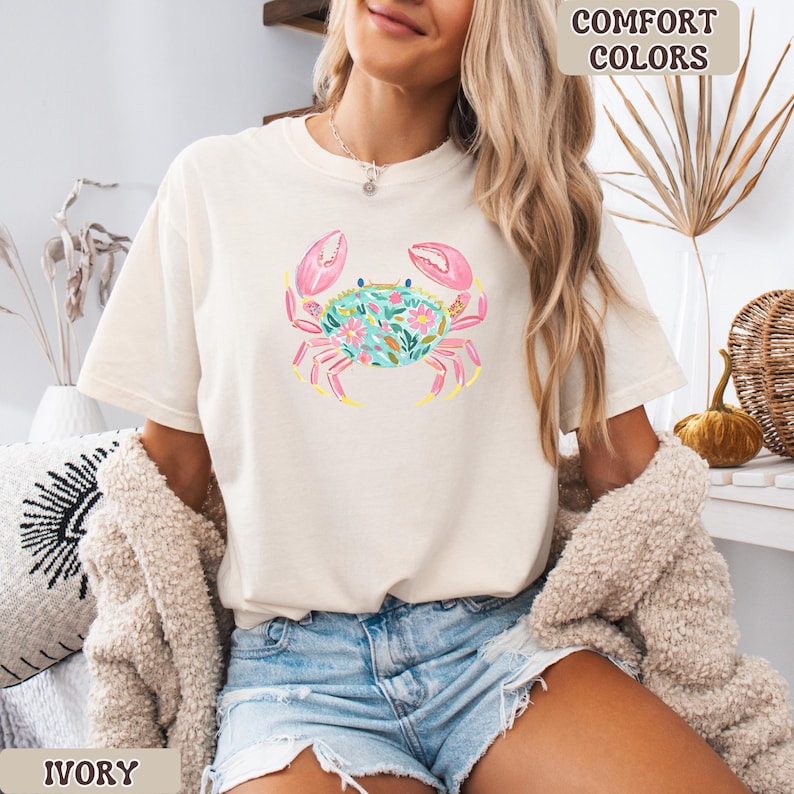 K&ouml;nnte beinhalten: Elfenbeinfarbenes T-Shirt mit einem Aquarell-Krebs-Design. Der Krebs hat rosa Scheren, einen t&uuml;rkisfarbenen K&ouml;rper voller Blumen und gelbe Akzente. Die Worte "Comfort Colors" und "Ivory" sind auf dem Bild zu sehen.