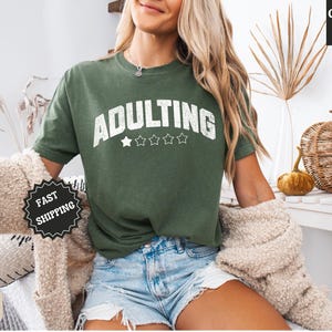 May include: Green t-shirt with the word "ADULTING" in white, above five white stars. The shirt is a Comfort Colors brand. The model is wearing denim shorts and a beige sweater. The image also includes a pumpkin and a decorative basket.