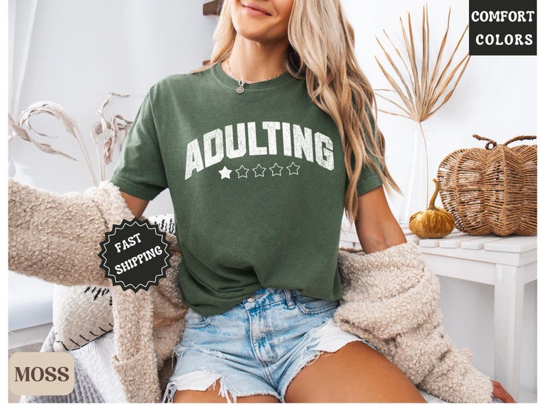 May include: Green t-shirt with the word "ADULTING" in white, above five white stars. The shirt is a Comfort Colors brand. The model is wearing denim shorts and a beige sweater. The image also includes a pumpkin and a decorative basket.