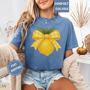 Puede incluir: Una camiseta azul vaquero con un lim&oacute;n amarillo y un lazo de cuadros amarillos y blancos. El texto "COMFORT COLORS" est&aacute; en la esquina superior derecha de la imagen.