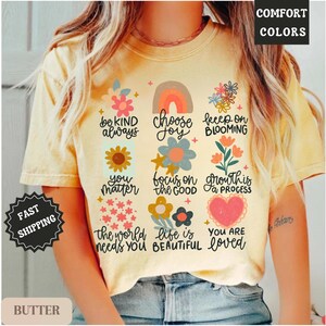 Pu&ograve; includere: Una maglietta gialla con una variet&agrave; di grafiche e testi colorati. Il testo include "be kind always", "choose joy", "keep on blooming", "you matter", "focus on the good", "growth is a process", "the world needs you", "life is beautiful" e "you are loved".