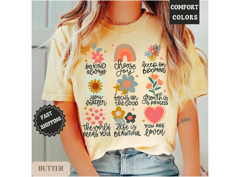 Pu&ograve; includere: Una maglietta gialla con una variet&agrave; di grafiche e testi colorati. Il testo include "be kind always", "choose joy", "keep on blooming", "you matter", "focus on the good", "growth is a process", "the world needs you", "life is beautiful" e "you are loved".