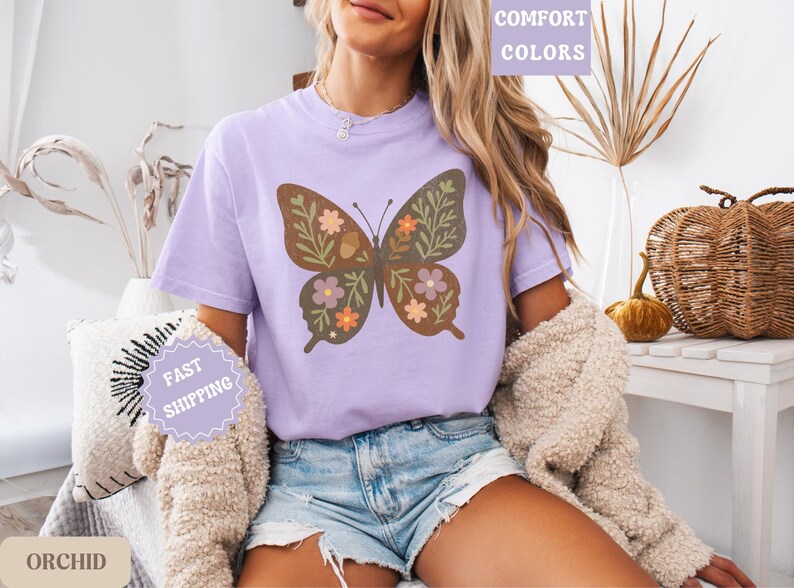 Peut inclure: T-shirt lilas avec un motif de papillon orn&eacute; de fleurs et de feuilles marron, orange et violet. Fait partie de la collection "Comfort Colors" et est &eacute;tiquet&eacute; "Orchid" avec un message "Livraison rapide".