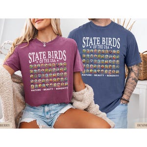 May include: Two t-shirts with a graphic design of state birds of the USA. The text on the shirts reads "State Birds of the USA" and "Nature - Beauty - Serenity". The shirt on the left is a berry color and the shirt on the right is denim blue.