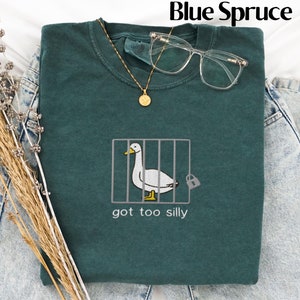 May include: A blue spruce sweatshirt with a humorous embroidered design of a duck in a cage, accompanied by the text "got too silly." The sweatshirt is styled with glasses and a gold necklace, and is laid on top of denim jeans.
