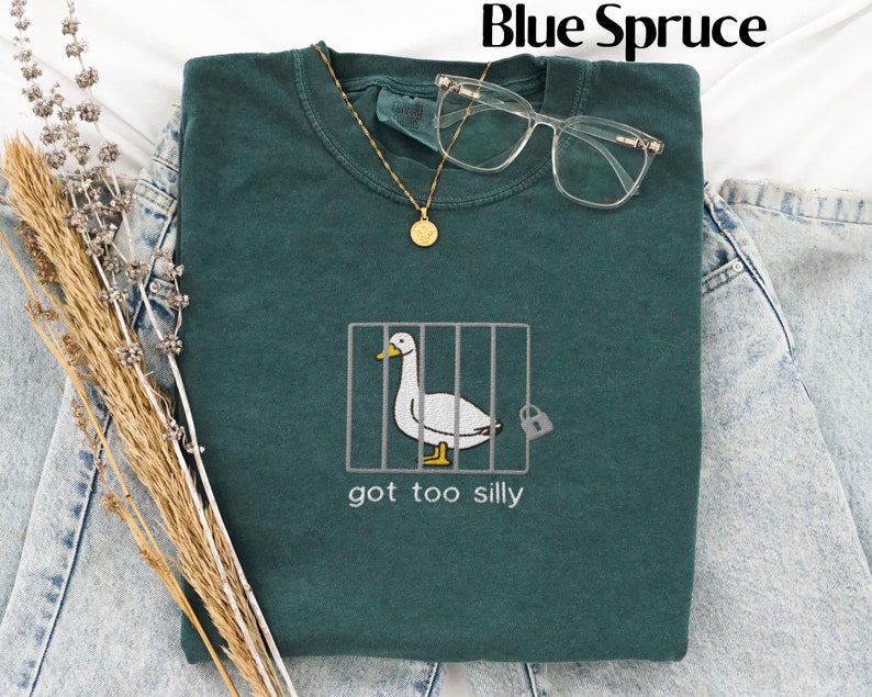 May include: A blue spruce sweatshirt with a humorous embroidered design of a duck in a cage, accompanied by the text "got too silly." The sweatshirt is styled with glasses and a gold necklace, and is laid on top of denim jeans.