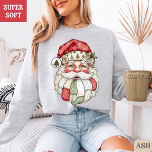 May include: Pale grey sweatshirt with a festive Santa Claus face design. The Santa face has a red hat, white beard, and a patchwork design on the beard and coat. The text "SUPER SOFT" is in a red box in the upper left corner.