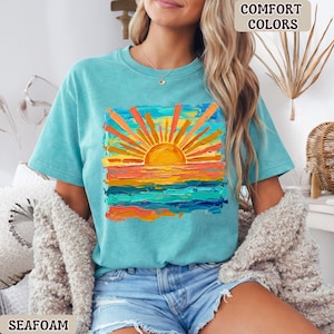 Puede incluir: Camiseta verde mar con un vibrante gr&aacute;fico de puesta de sol. La obra de arte presenta un sol naranja brillante con rayos radiantes sobre un horizonte colorido de naranja, amarillo y azul. Las palabras "COMFORT COLORS" est&aacute;n impresas en la parte superior.