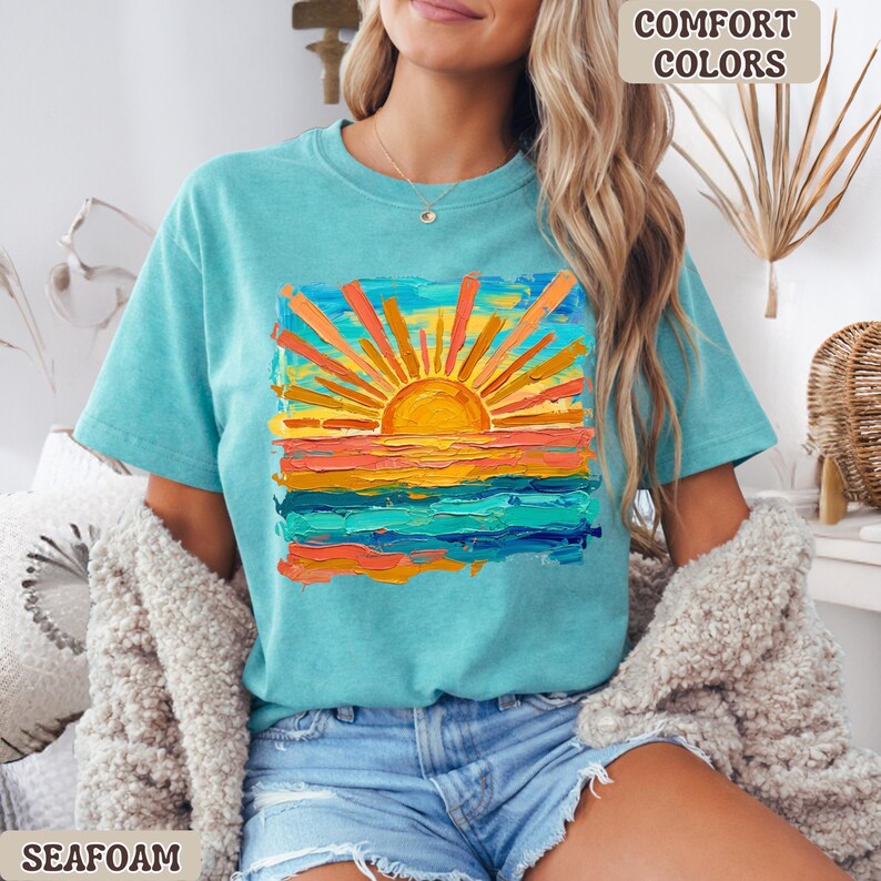 Puede incluir: Camiseta verde mar con un vibrante gr&aacute;fico de puesta de sol. La obra de arte presenta un sol naranja brillante con rayos radiantes sobre un horizonte colorido de naranja, amarillo y azul. Las palabras "COMFORT COLORS" est&aacute;n impresas en la parte superior.