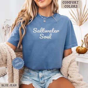 Op de afbeelding: Een blauw spijkerbroekkleurig t-shirt met de woorden "Saltwater Soul" in witte letters. Het shirt heeft korte mouwen en lijkt van een zacht materiaal te zijn. De woorden "Comfort Colors" staan in een bruine doos rechtsboven.